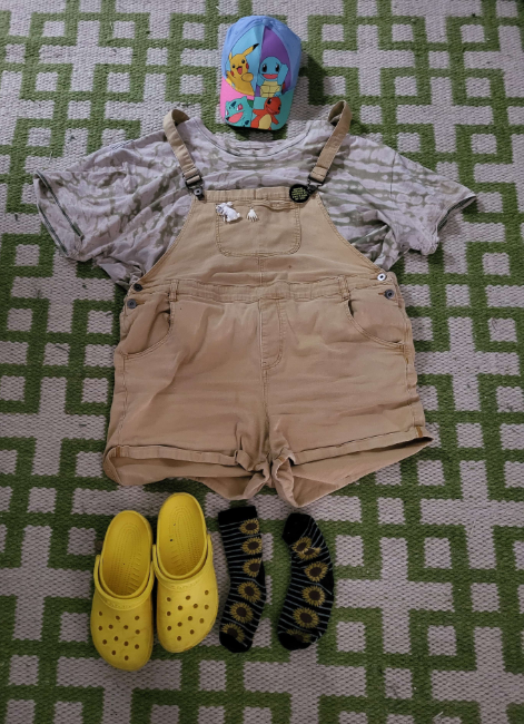 a bleach tye dyed green shirt a pair of yellow overall shorts a pokemon baseball cap an earring a rabbit pin a circular pin sunflower socks and yellow crocs placed over a green and white geometric patterened rug