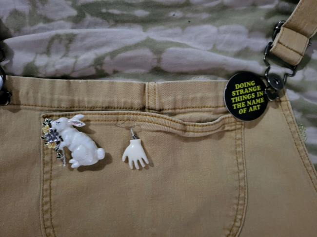 a closeup of the rabbit pin the white hand earring and the lush pin that reads do strange things in the name of art